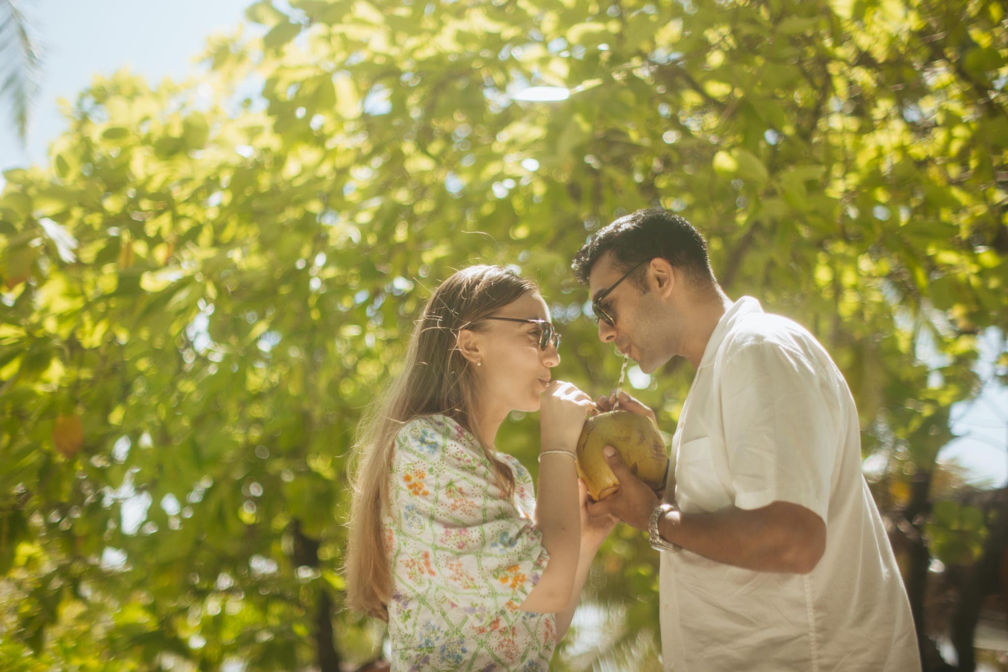 Couple sharing coconuts under sunlit green tree canopy