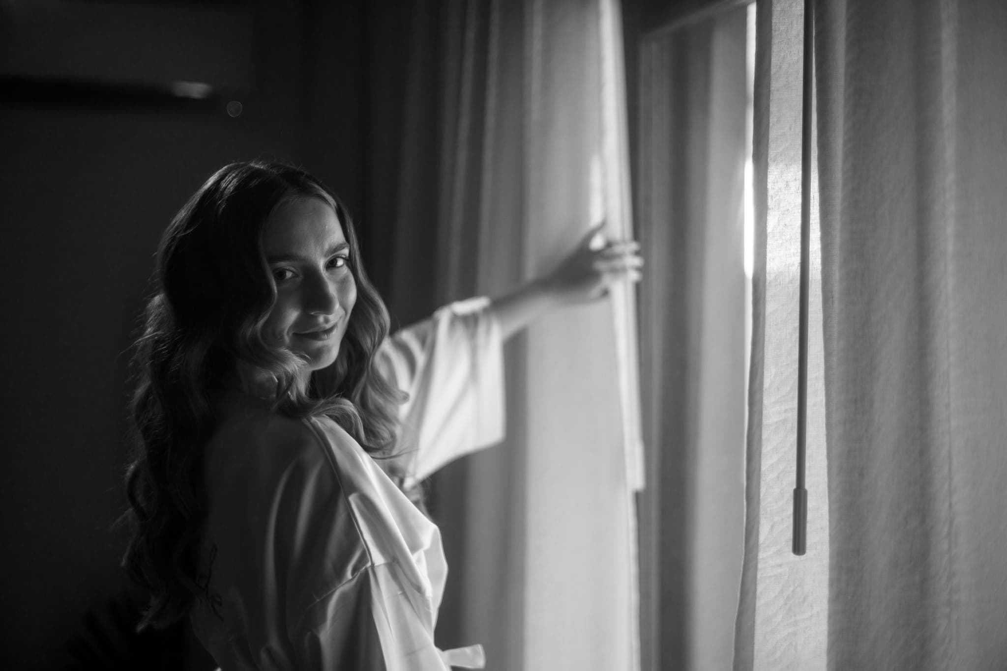 Bride glancing back while opening curtains in soft light