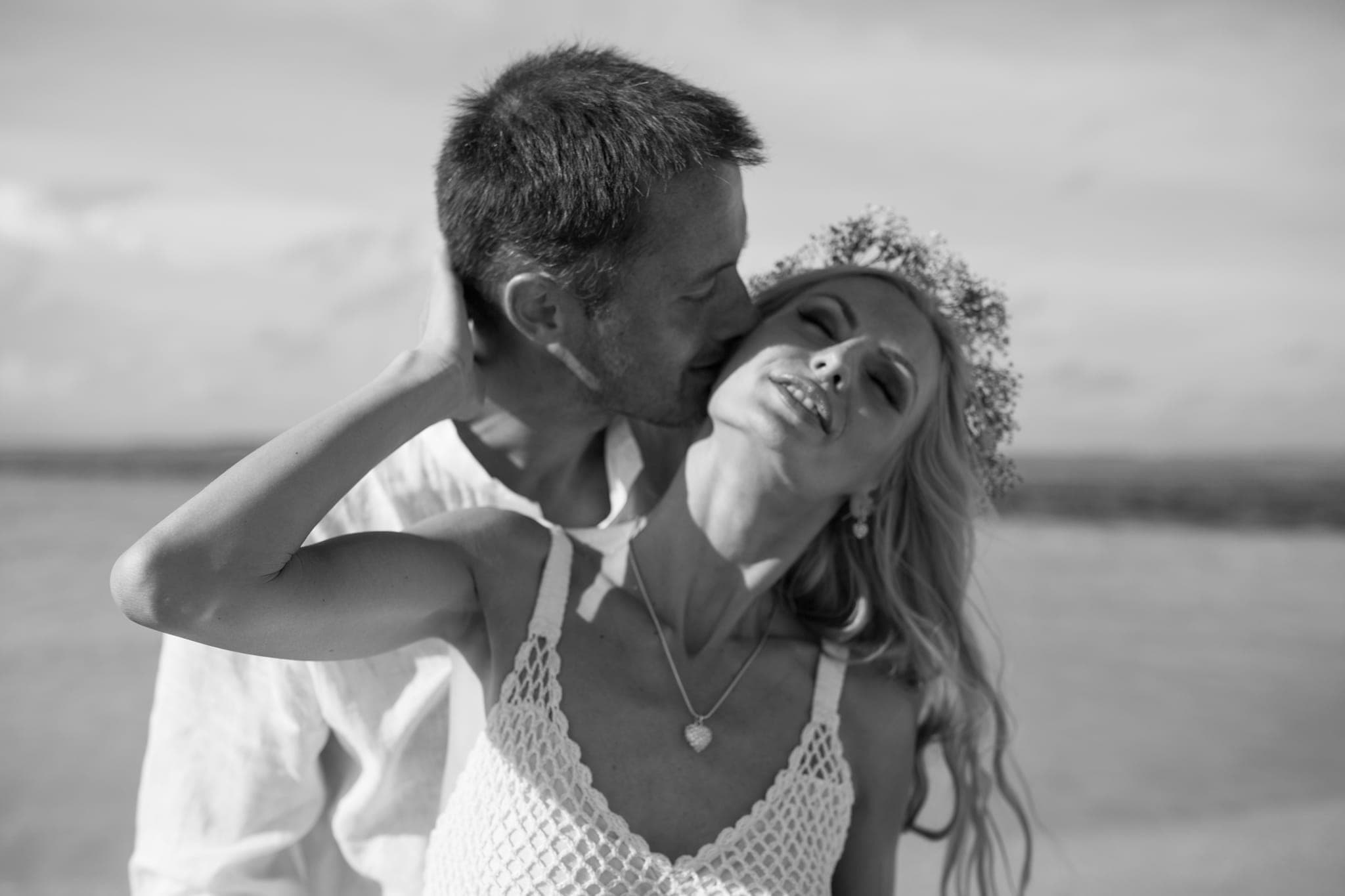 Groom kissing bride's forehead on beach, black and white
