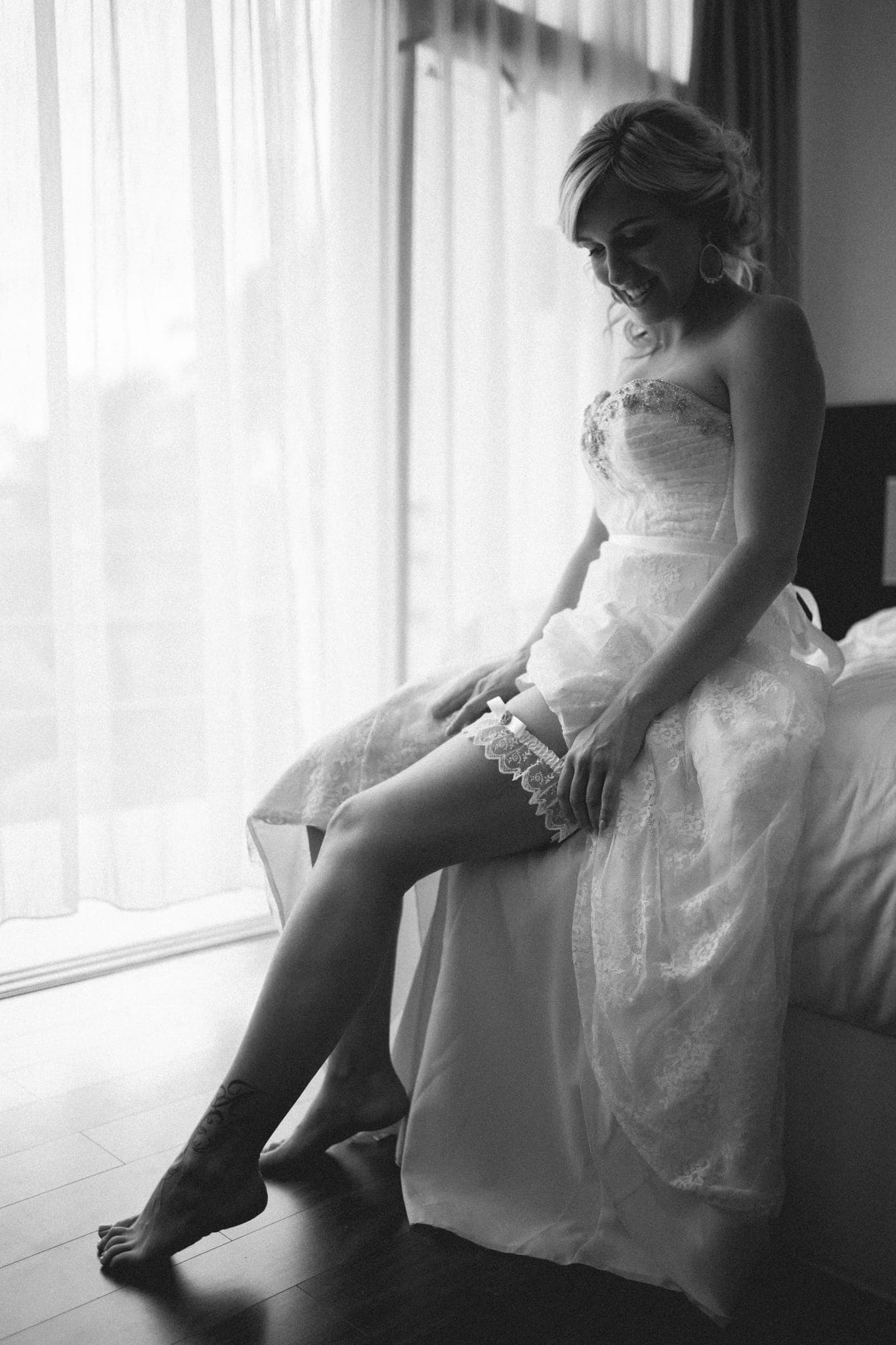 Bride sitting on bed by window putting on garter
