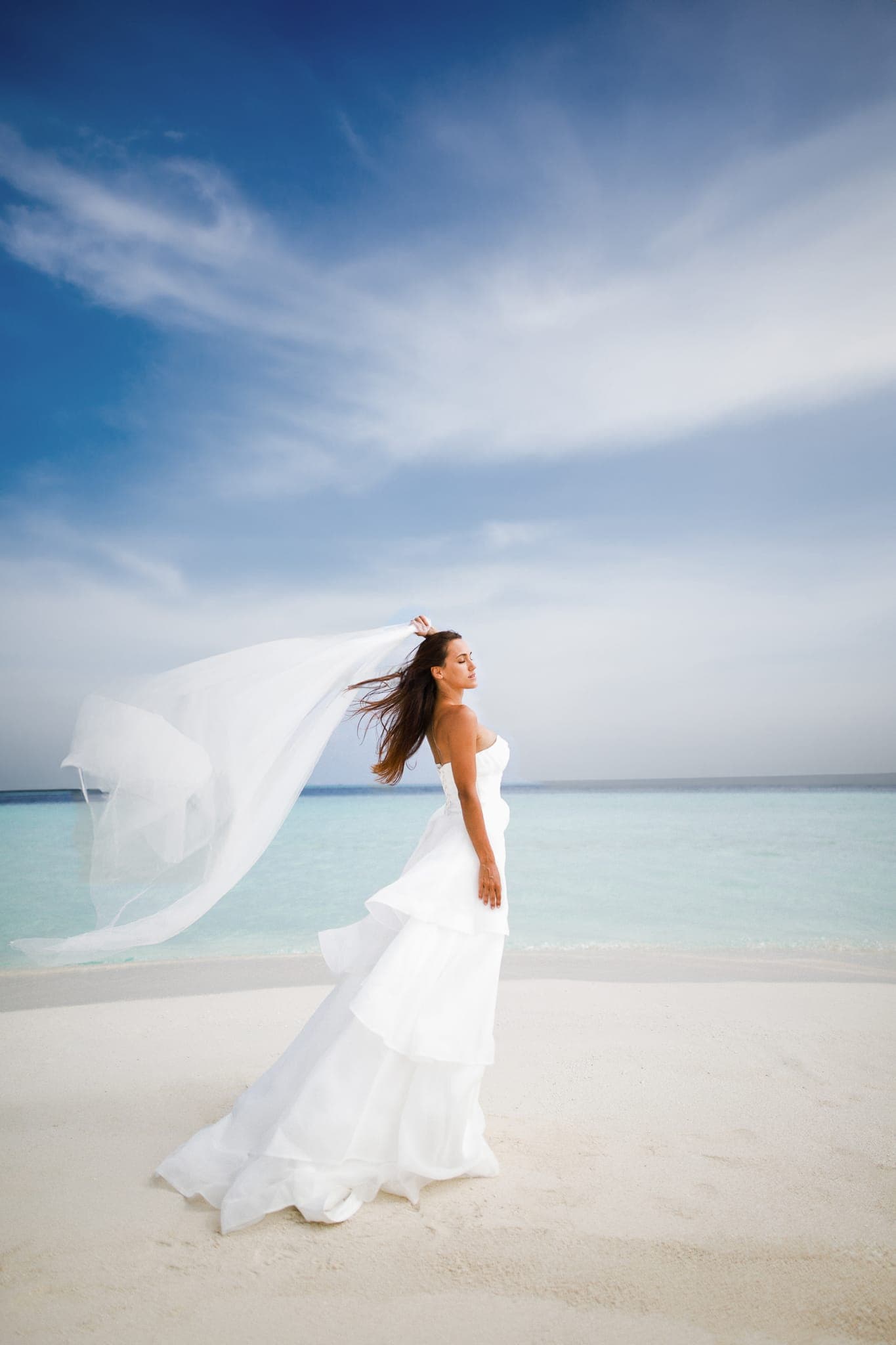 Bride in white gown, veil flowing on tropical beach