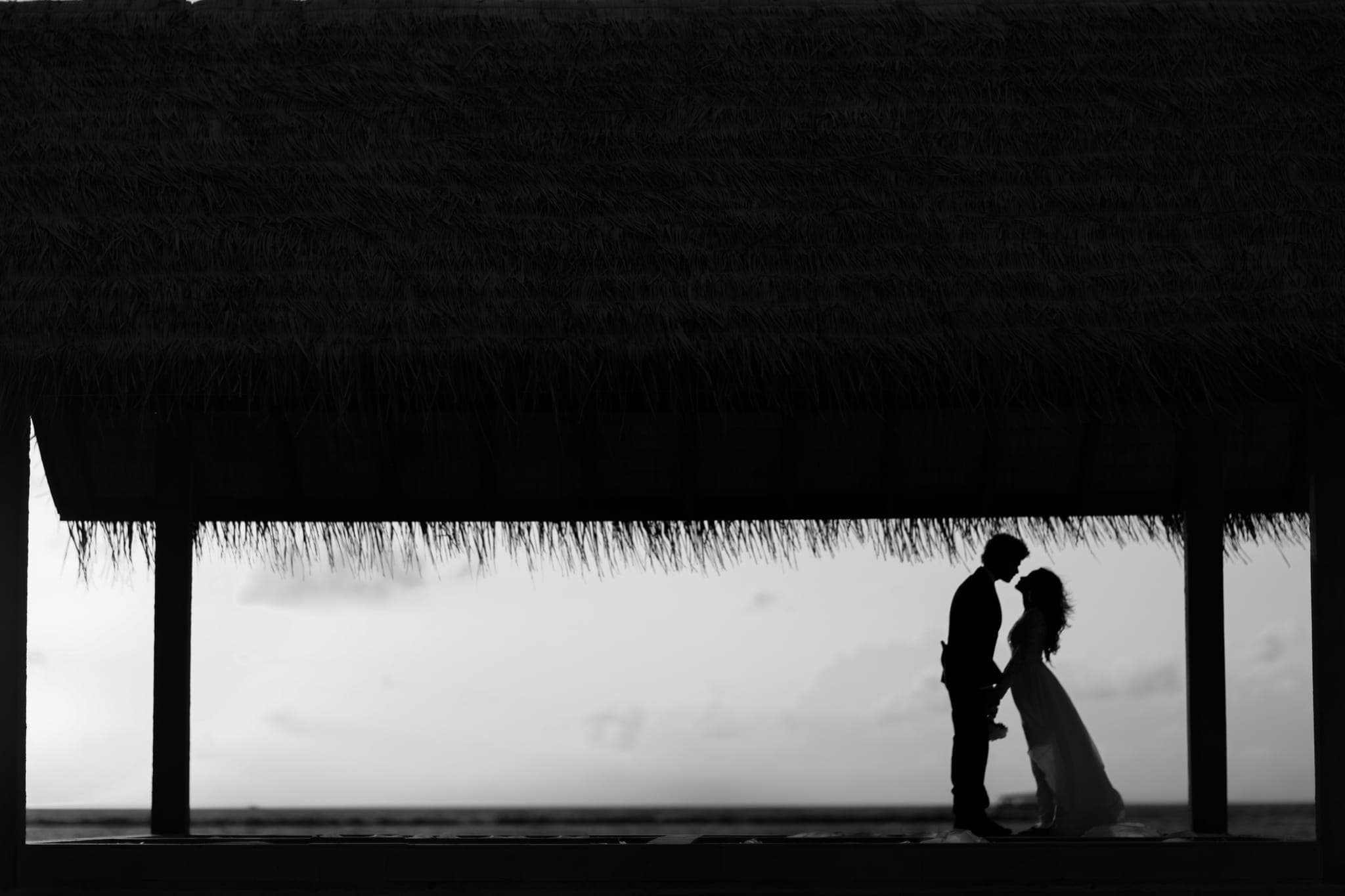 Silhouetted couple kissing under thatched roof by ocean