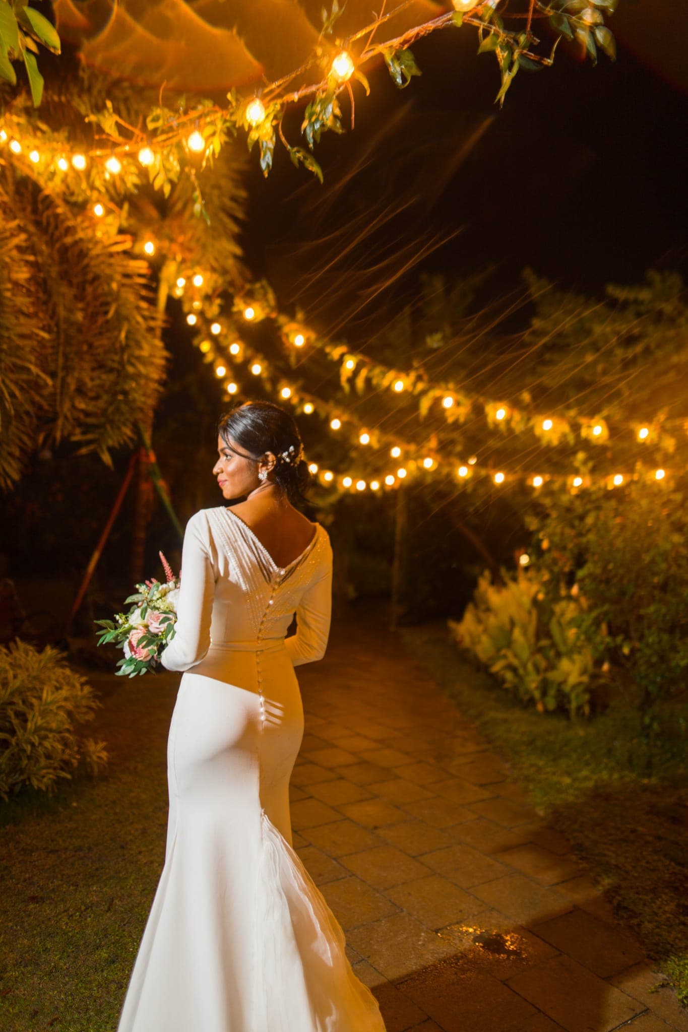 Bride walking garden path under glowing string lights