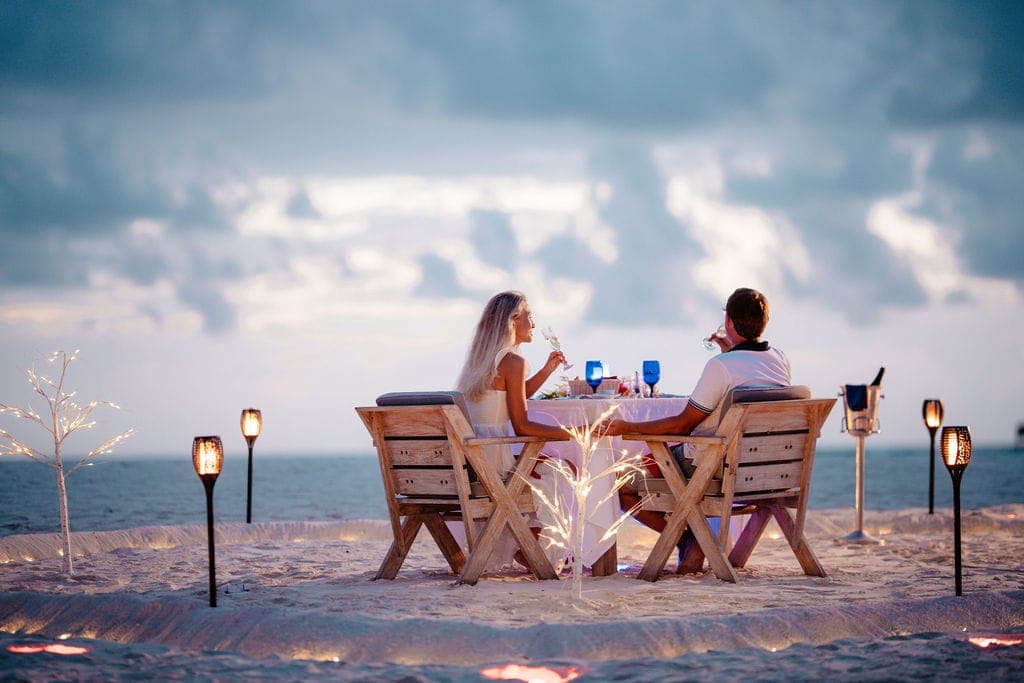 Couple at candlelit beach dinner table under dusk sky