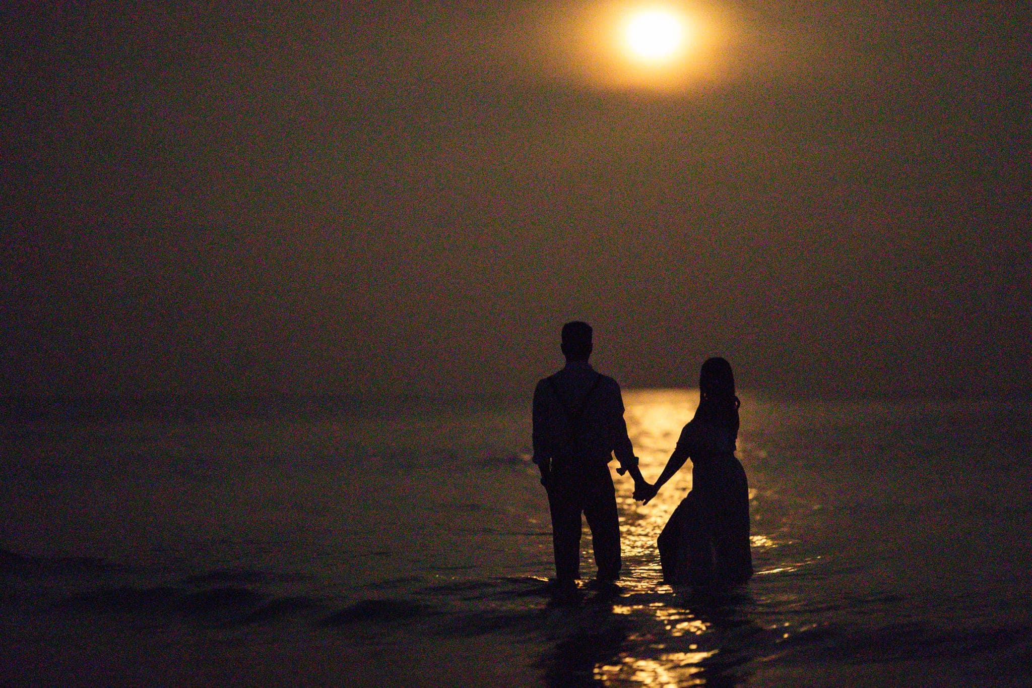 Couple silhouetted holding hands in ocean under moonlight
