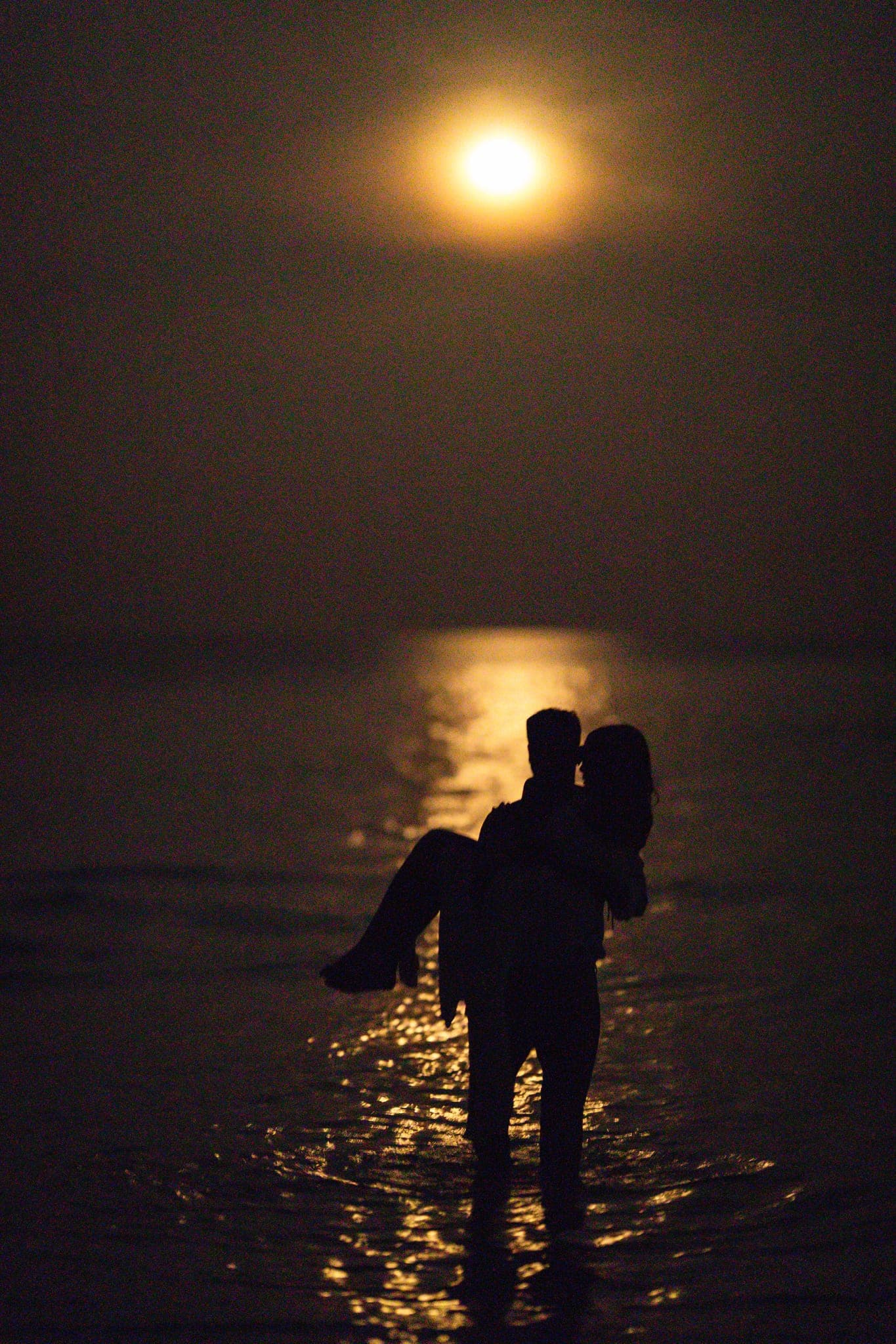 Groom carrying bride through moonlit ocean water