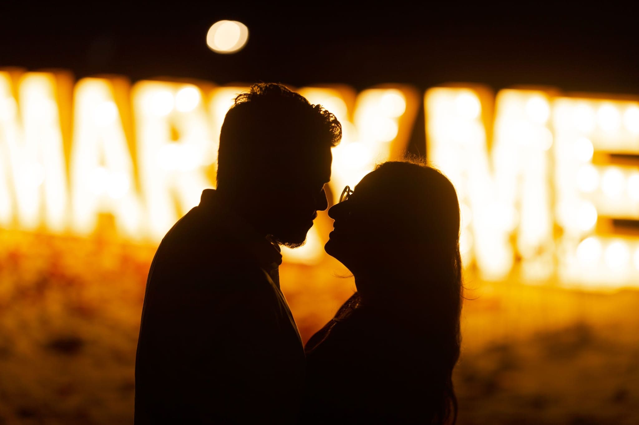 Couple silhouetted kissing beside glowing marry me sign