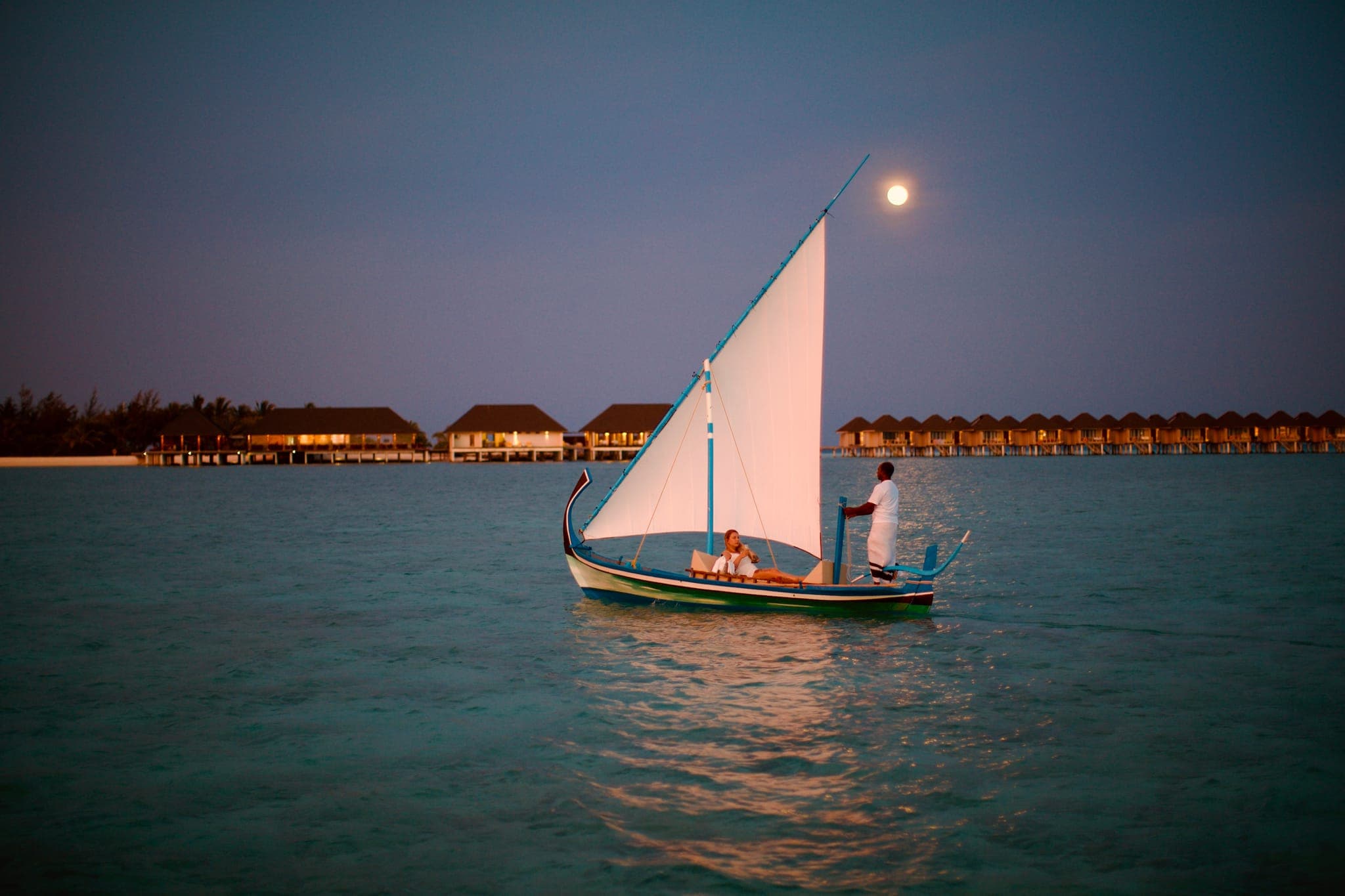 Sailboat crossing moonlit lagoon past water villas