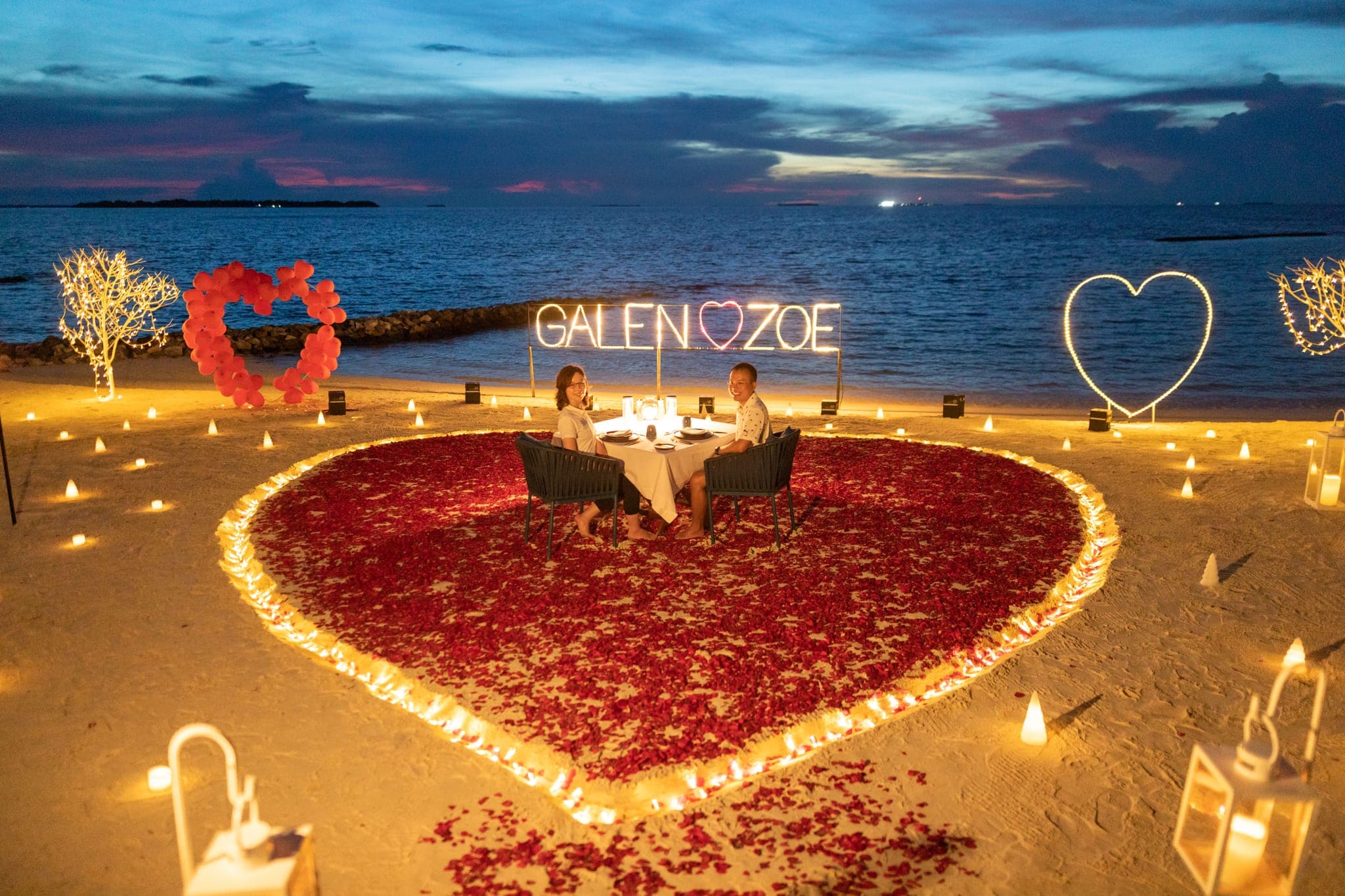 Couple at candlelit heart-shaped beach proposal setup at dusk